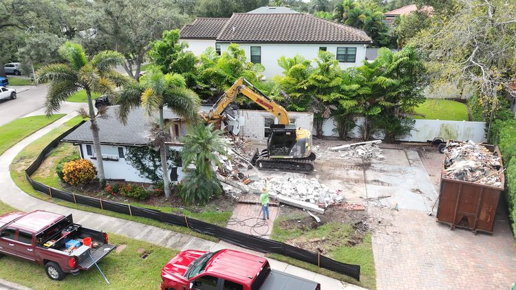 Aerial view of a Davis Islands lot mid-demolition, with an excavator, a full dumpster and silt fence along the sidewalk