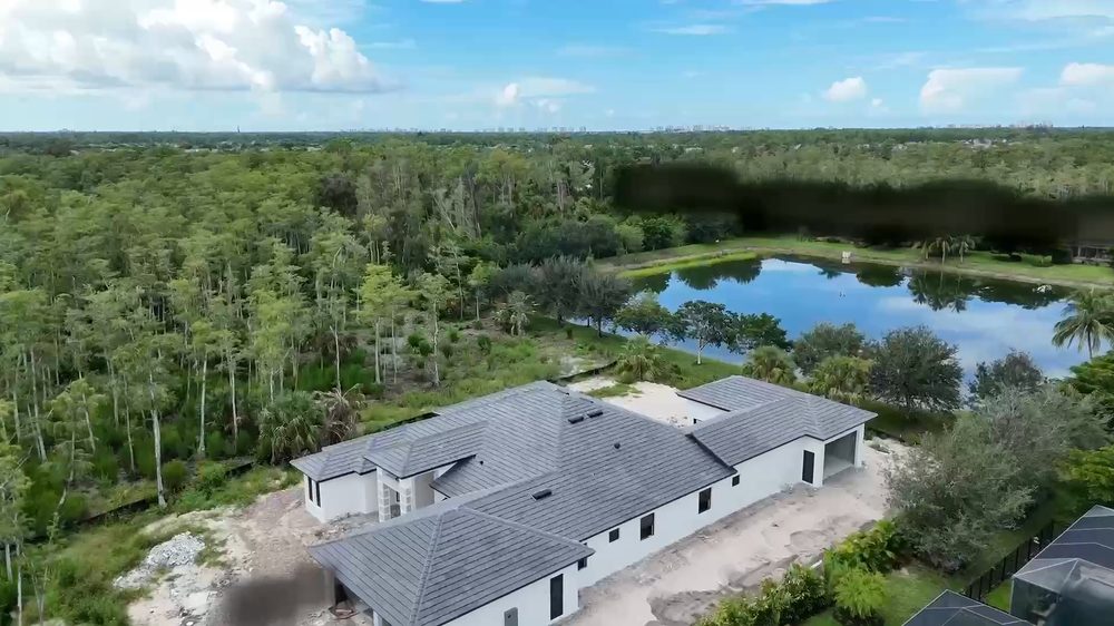 Aerial view of a single-story custom home with a gray tile roof on a wooded Naples lot near a pond